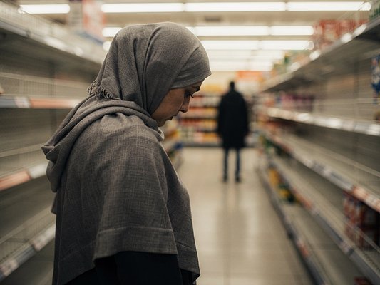 Una donna con il velo tra gli scaffali di un supermercato, seguita da una figura scura in fondo alla corsia.